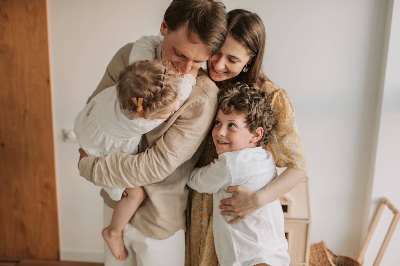 Two adults and two children standing indoors, sharing a group hug