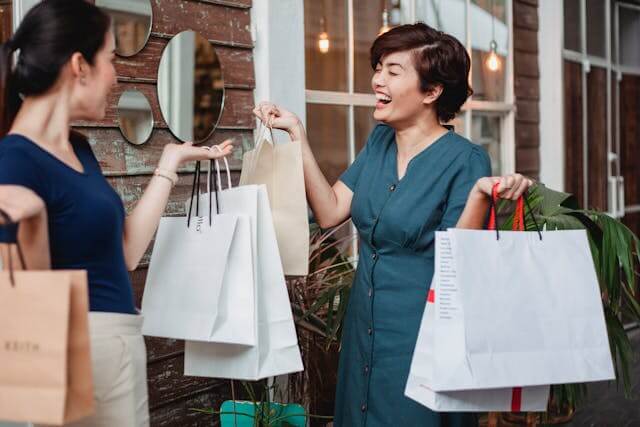 Two women standing outside with shopping bags, smiling and showing each other their purchases.