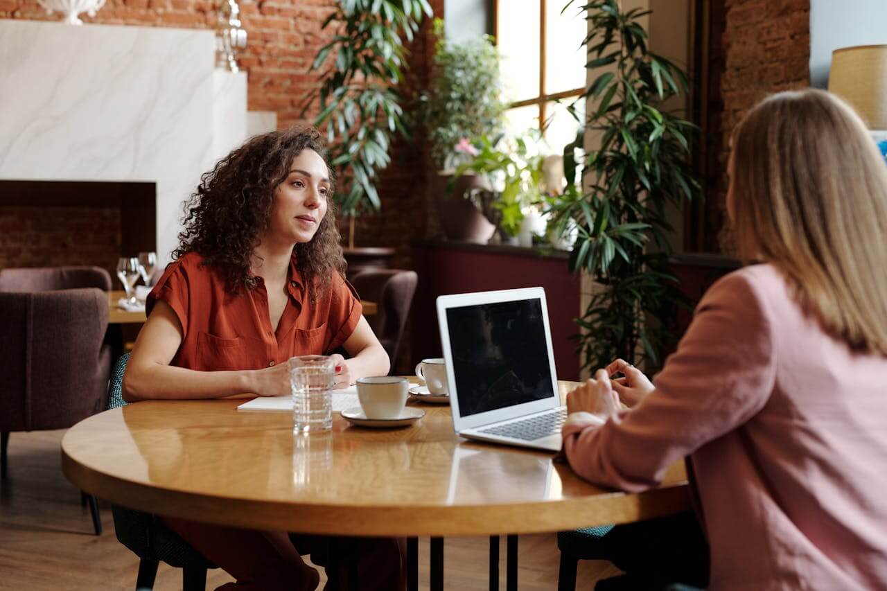 Two people sitting at a table having a conversation, with a laptop open between them