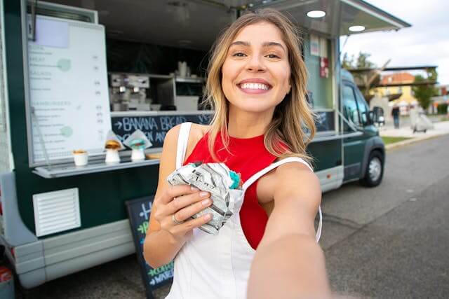 A person smiling and holding a drink while standing in front of a food truck outdoors.