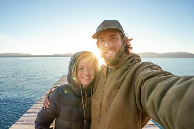 Couple taking a selfie together outdoors by the water, smiling in relaxed clothing.