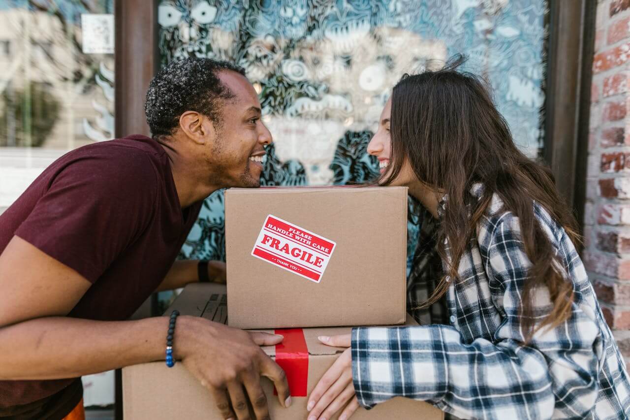 Two people smiling while holding a cardboard moving box outside a home.