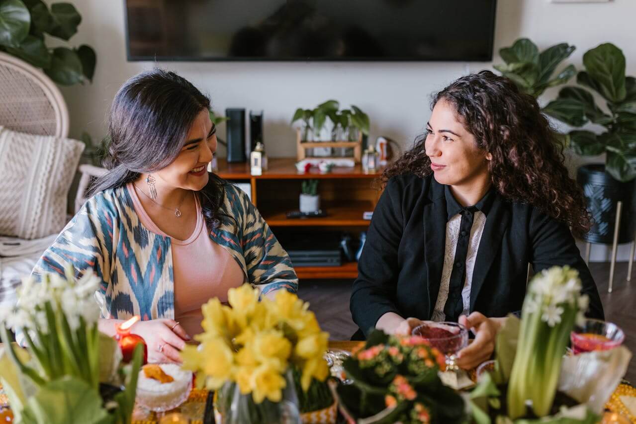 Two women sitting at a table at home, talking and smiling, with flowers arranged in the foreground.